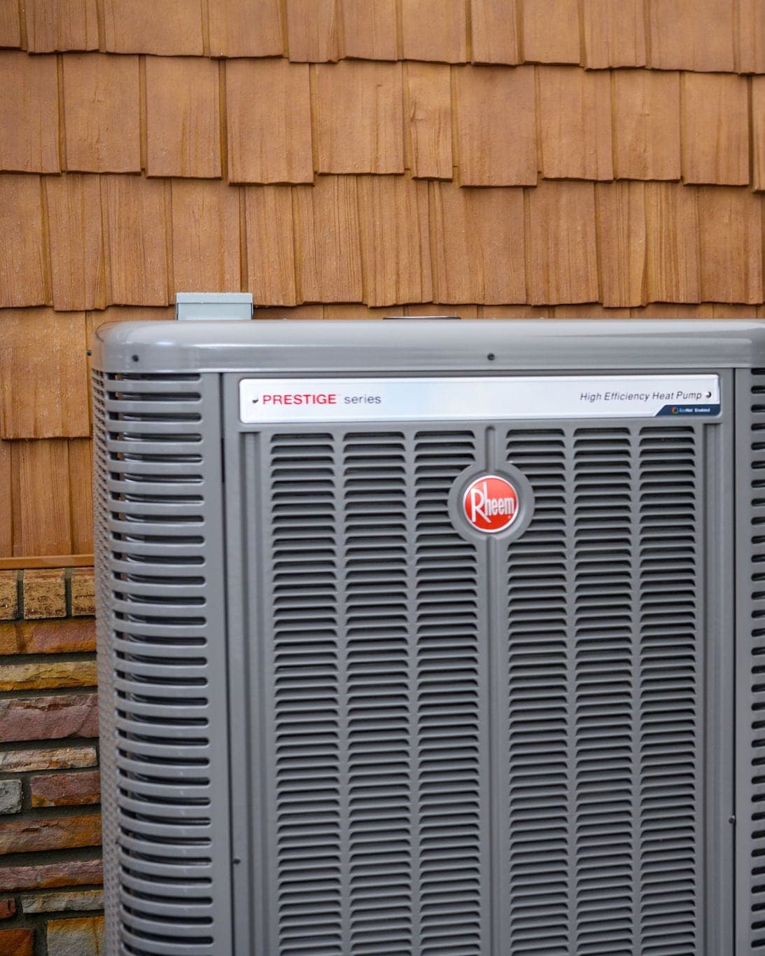 A Rheem Prestige series high efficiency heat pump installed beside a shingled house wall.